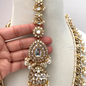 May include: A gold and white Indian bridal necklace set with pearl and kundan embellishments. The necklace features a long, layered design with multiple strands of pearls and kundan stones. The set also includes a matching maang tikka, a traditional Indian headpiece.
