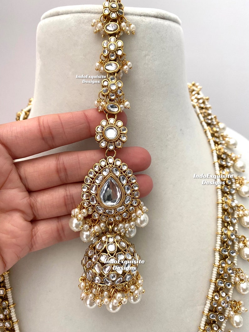 May include: A gold and white Indian bridal necklace set with pearl and kundan embellishments. The necklace features a long, layered design with multiple strands of pearls and kundan stones. The set also includes a matching maang tikka, a traditional Indian headpiece.