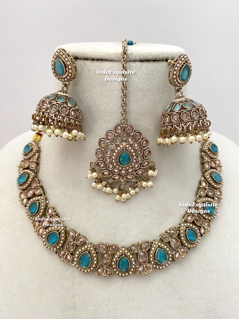May include: An antique-style jewelry set featuring a necklace, earrings, and a headpiece. The set has a bronze finish with teal-colored teardrop-shaped stones and small white pearls. The earrings are bell-shaped. The text "IndoExquisite Designs" is visible.
