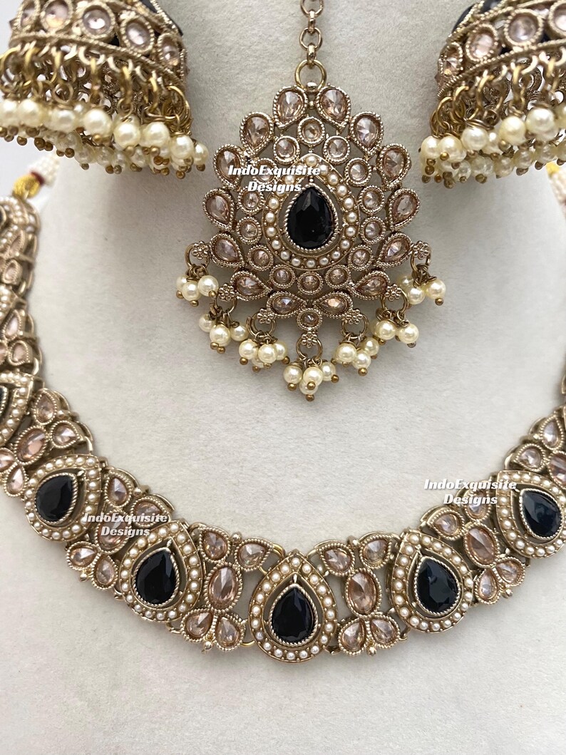 May include: An ornate jewelry set featuring a necklace, earrings, and a headpiece. The set is gold-toned with black teardrop-shaped stones and small white pearls. The necklace and headpiece have a floral design.