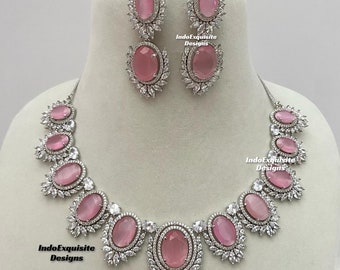 May include: A silver necklace and earrings set with pink oval stones and clear cubic zirconia accents. The necklace features a row of stones with smaller stones surrounding each stone. The earrings are drop earrings with two stones.