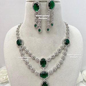 Puede incluir: Un conjunto de collar y pendientes plateados con grandes piedras preciosas de color verde esmeralda de forma ovalada y de pera. El collar tiene dos hileras con piedras claras más pequeñas. Los pendientes tienen un diseño en cascada.