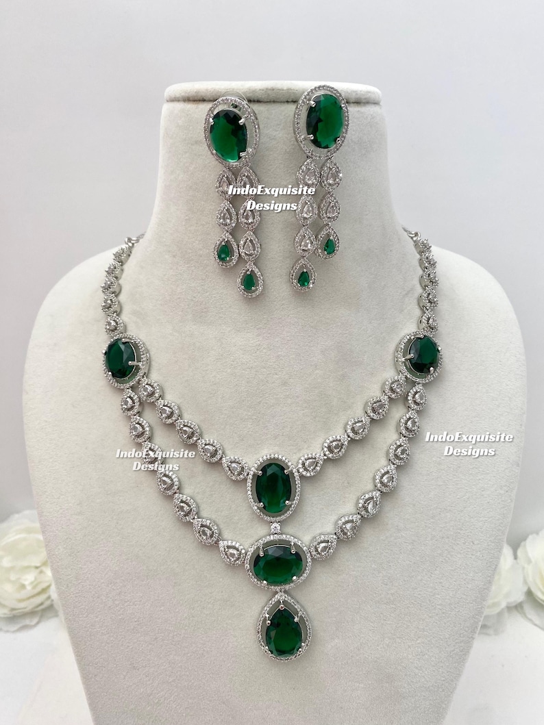 May include: A silver-toned necklace and earring set featuring large oval and pear-shaped emerald green gemstones. The necklace has two strands with smaller clear stones. The earrings have a cascading design with clear and green stones.