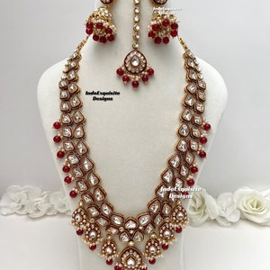 Puede incluir: Un conjunto de collar de novia indio en oro y rojo con un collar largo de varias capas, pendientes a juego y un tikka. El collar presenta diseños intrincados con piedras blancas y cuentas rojas.