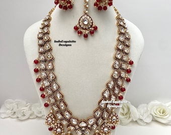 Op de afbeelding: Een gouden en rode Indiase bruids kettingset met een lange, gelaagde ketting, bijpassende oorbellen en een tikka. De ketting heeft ingewikkelde ontwerpen met witte stenen en rode kralen.