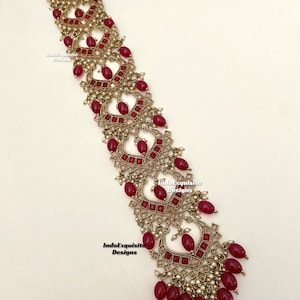 May include: A gold and red hair accessory with a decorative design featuring small red and white beads. The accessory is made of metal and has a traditional Indian design.