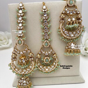 Puede incluir: Un conjunto de joyer&iacute;a en tono dorado que incluye dos pendientes grandes en forma de media luna y un maang tikka a juego. Cada pieza est&aacute; adornada con piedras transparentes, detalles en forma de gota verde claro y peque&ntilde;as cuentas verde claro. El texto IndoExquisite Designs es visible.