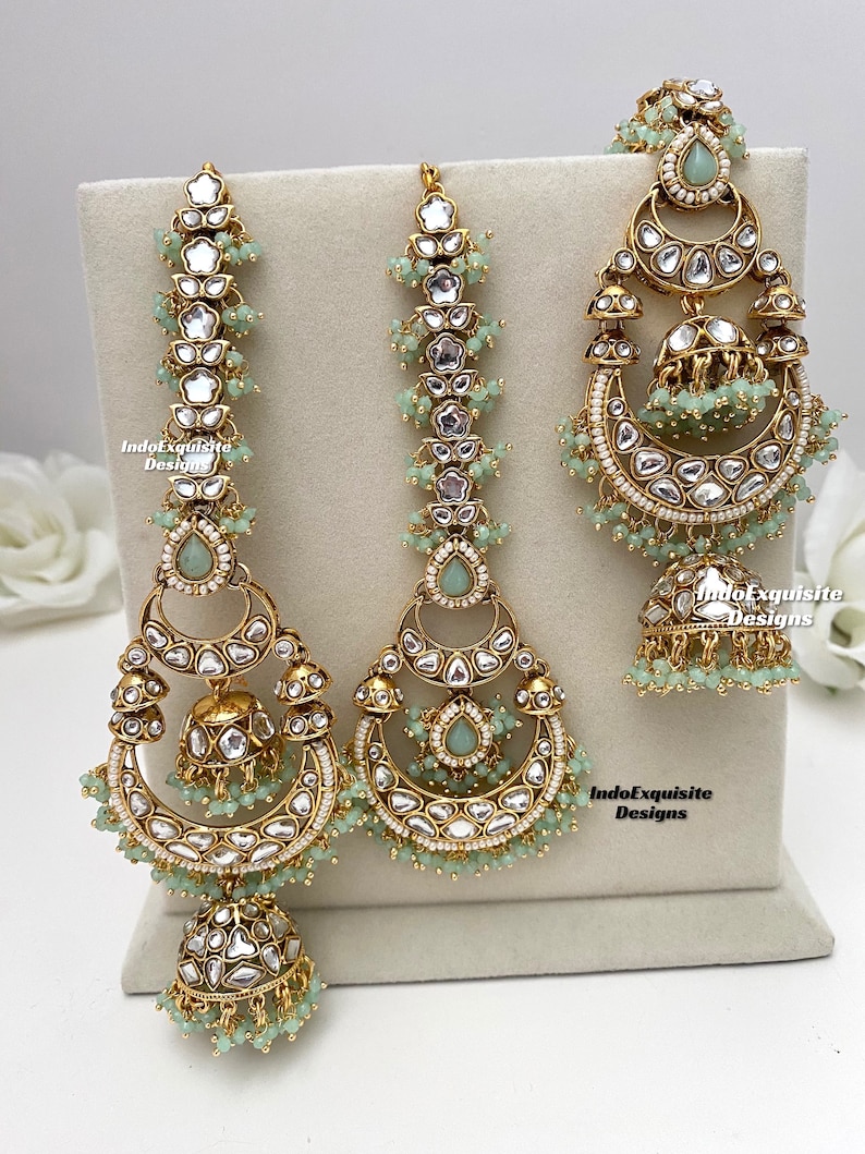 Puede incluir: Un conjunto de joyer&iacute;a en tono dorado que incluye dos pendientes grandes en forma de media luna y un maang tikka a juego. Cada pieza est&aacute; adornada con piedras transparentes, detalles en forma de gota verde claro y peque&ntilde;as cuentas verde claro. El texto IndoExquisite Designs es visible.