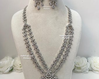 Puede incluir: Un conjunto de collar y pendientes en tono plateado con cristales transparentes y piedras azules en forma de lágrima. El collar tiene una cadena de doble hebra y un colgante grande y ornamentado. El texto IndoExquisite Designs es visible.