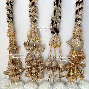 May include: Four ornate, gold-colored hair accessories with black braided cords. Each features intricate beadwork, dangling ornaments, and decorative elements. White fabric flowers are at the base of the accessories. The text "IndoExquisite Designs" is visible.