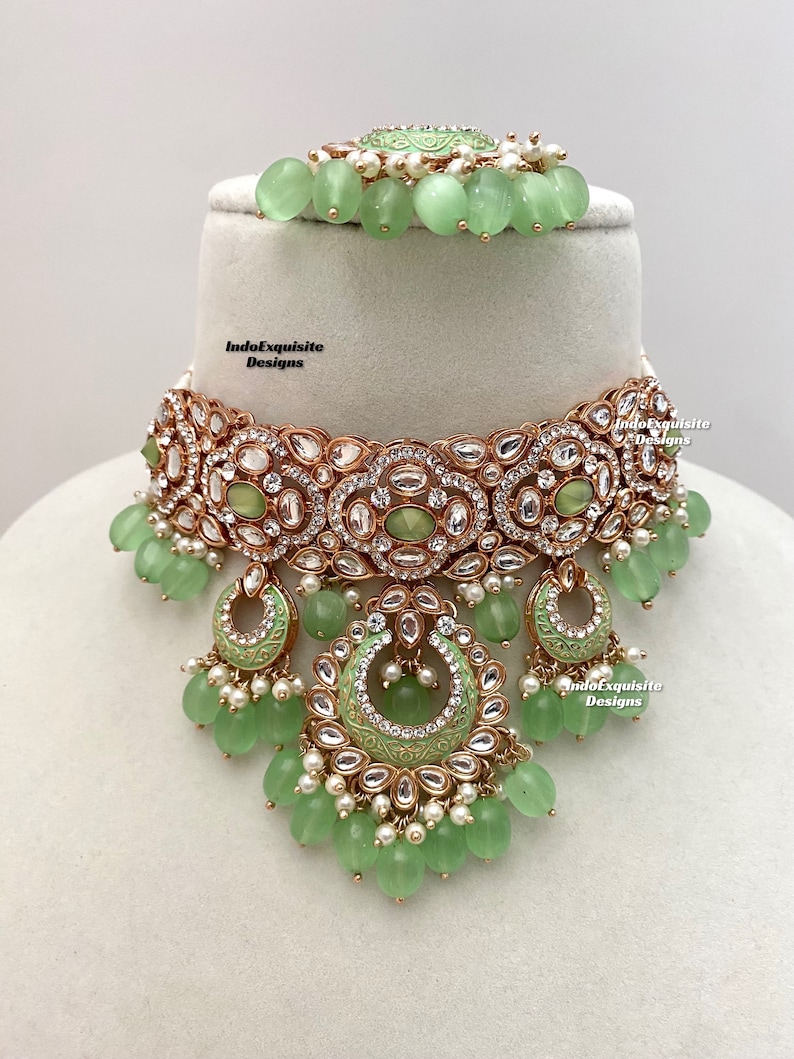 May include: A green and gold choker necklace with a large, ornate pendant. The pendant is decorated with pearls, rhinestones, and a green enamel design. The necklace is accented with green beads.