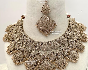 Puede incluir: Un conjunto de collar y maang tikka en tono dorado con intrincados trabajos de metal en filigrana, incrustado con piedras transparentes y acentuado con pequeñas cuentas colgantes. El conjunto de joyería muestra el texto IndoExquisite Designs.