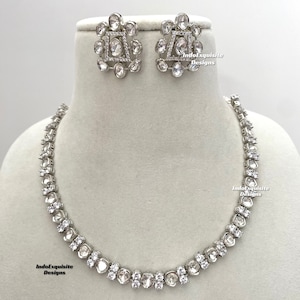 May include: A silver necklace with a row of round, clear stones. The necklace is on a white mannequin. There are matching earrings with a square design in the center and round stones around the square. The text "IndoExquisite Designs" is visible on the image.