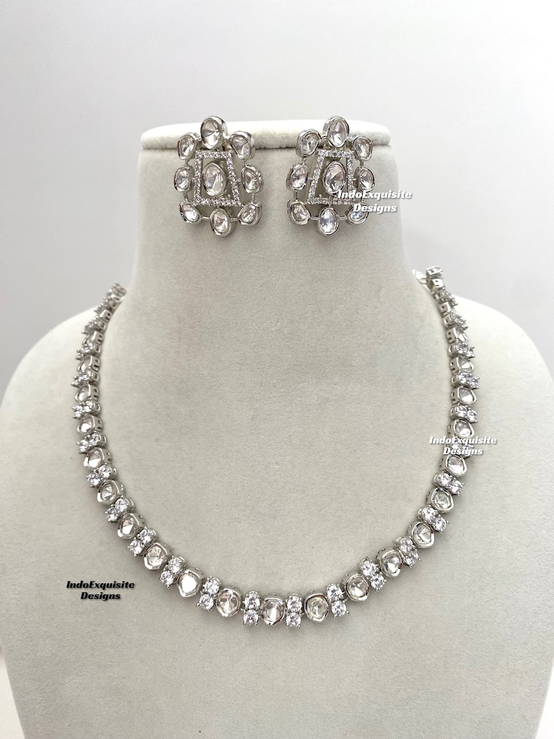 May include: A silver necklace with a row of round, clear stones. The necklace is on a white mannequin. There are matching earrings with a square design in the center and round stones around the square. The text "IndoExquisite Designs" is visible on the image.