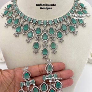 May include: A silver necklace and earrings set with teardrop-shaped green stones and clear rhinestones. The necklace has a layered design with multiple rows of stones. The earrings have a similar design with three teardrop-shaped stones.