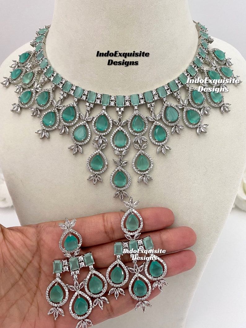 May include: A silver necklace and earrings set with teardrop-shaped green stones and clear rhinestones. The necklace has a layered design with multiple rows of stones. The earrings have a similar design with three teardrop-shaped stones.
