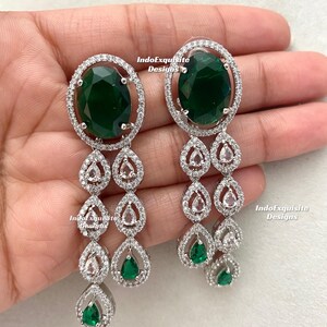 May include: Elegant dangle earrings featuring large oval emerald green stones surrounded by a halo of clear crystals. The earrings have a cascading design with teardrop-shaped accents, also adorned with crystals and smaller green stones. The text "IndoExquisite Designs" is visible.