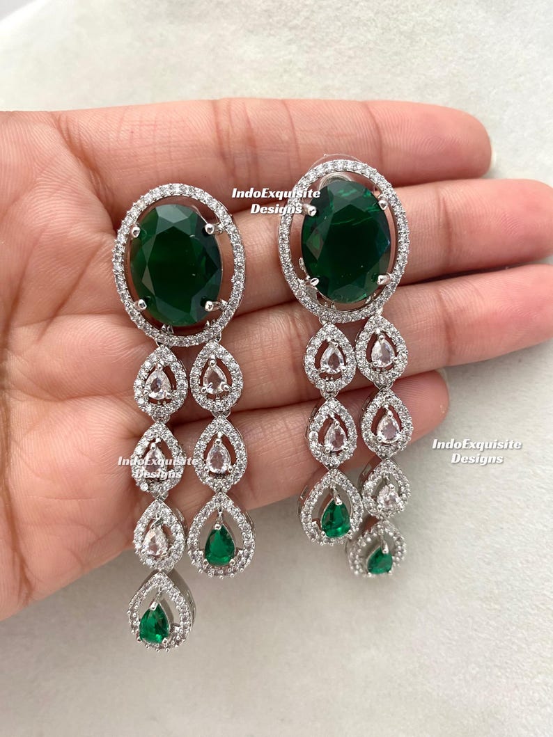 May include: Elegant dangle earrings featuring large oval emerald green stones surrounded by a halo of clear crystals. The earrings have a cascading design with teardrop-shaped accents, also adorned with crystals and smaller green stones. The text "IndoExquisite Designs" is visible.