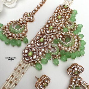 May include: A green and gold Indian bridal necklace set with a matching pair of earrings. The necklace features a choker style with a layered pearl chain and a large pendant with green stones and gold accents. The earrings are also adorned with green stones and gold accents. The set is perfect for a traditional Indian wedding or other special occasion.