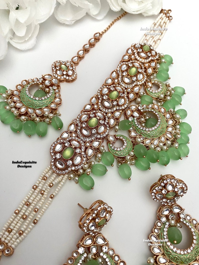 May include: A green and gold Indian bridal necklace set with a matching pair of earrings. The necklace features a choker style with a layered pearl chain and a large pendant with green stones and gold accents. The earrings are also adorned with green stones and gold accents. The set is perfect for a traditional Indian wedding or other special occasion.