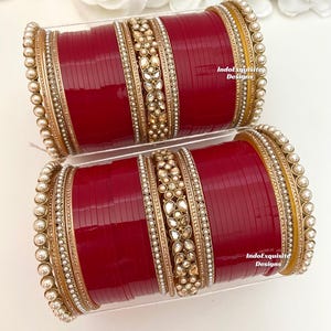 Può includere: Un set di braccialetti rossi e dorati con decorazioni di perle e strass. I braccialetti sono impilati uno sopra l'altro e disposti in un motivo circolare. I braccialetti sono decorati con disegni intricati e sono un accessorio tradizionale indiano.