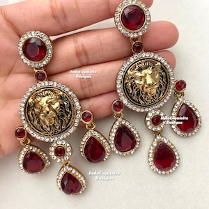May include: A pair of gold-tone earrings with a lion head design and red and clear crystal accents. The earrings are dangling and have a drop shape.