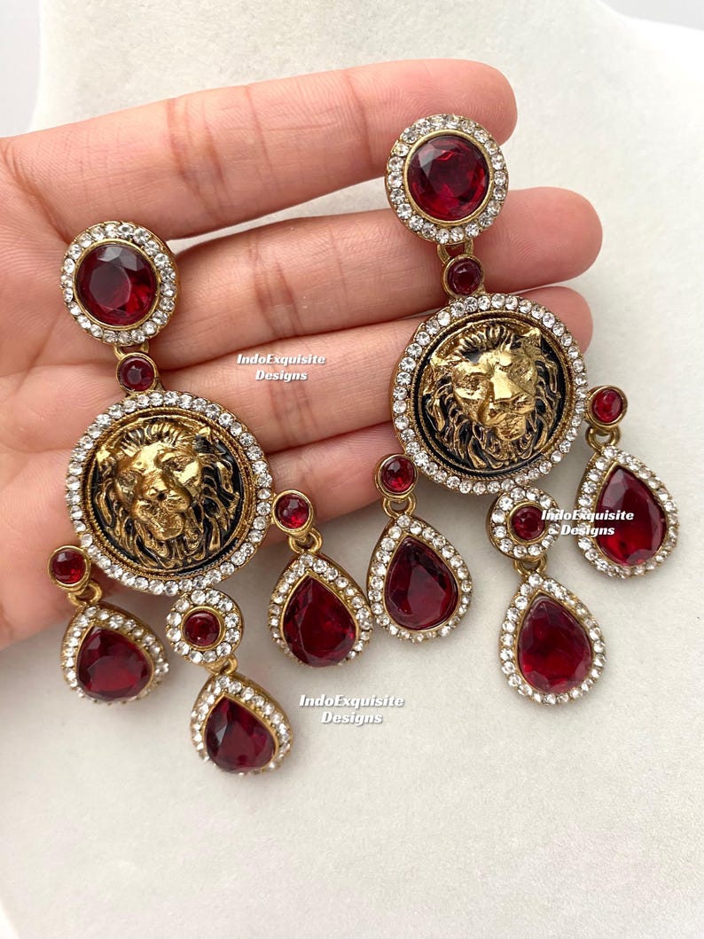 May include: A pair of gold-tone earrings with a lion head design and red and clear crystal accents. The earrings are dangling and have a drop shape.