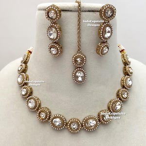 May include: A gold-coloured jewellery set comprising a necklace, earrings, and a headpiece. The set features oval-shaped clear stones encircled by small white pearls. The text "IndoExquisite Designs" is visible on the jewellery.