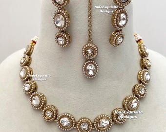 Puede incluir: Un conjunto de joyería en tono dorado que incluye un collar, dos pendientes colgantes y un maang tikka. Cada pieza contiene piedras ovaladas transparentes rodeadas de pequeñas perlas blancas. El texto IndoExquisite Designs es visible.