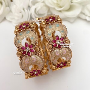 May include: Two gold bangles with intricate white and pink enamel designs. The bangles are decorated with pink and white gemstones and have a floral motif.