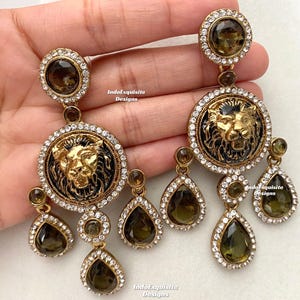 May include: A pair of gold-tone earrings with a lion head design and teardrop-shaped crystals. The lion head is surrounded by clear rhinestones and the teardrop crystals are a dark green color.