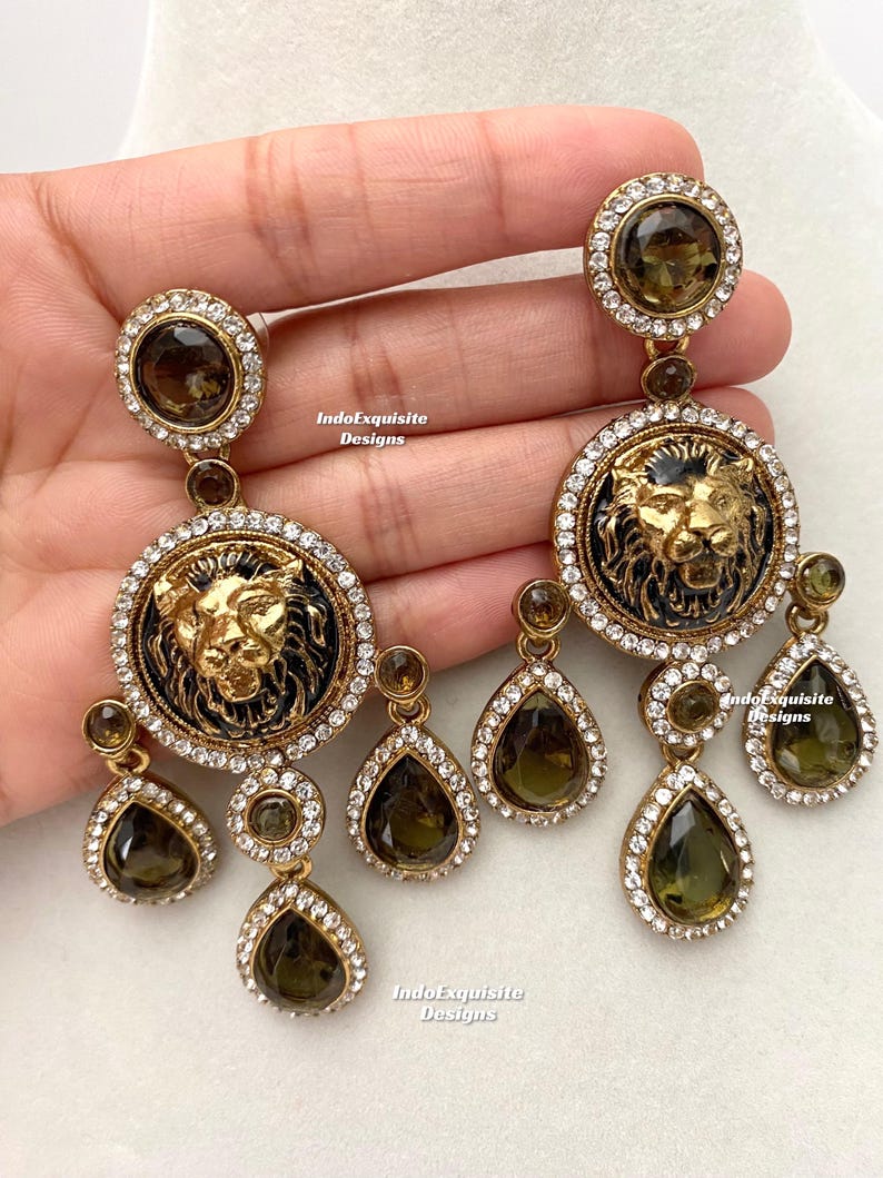 May include: A pair of gold-tone earrings with a lion head design and teardrop-shaped crystals. The lion head is surrounded by clear rhinestones and the teardrop crystals are a dark green color.
