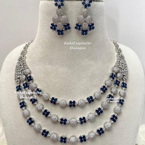 May include: A three-strand necklace and matching earrings set. The necklace features silver-colored beads and dark blue flower-shaped accents. The earrings have a similar design, with silver-colored beads and dark blue flower accents. The text "IndoExquisite Designs" is visible.