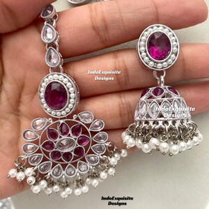 Op de afbeelding: Een zilveren ketting en oorbellen set met roze en witte stenen. De ketting heeft een hanger met een rond ontwerp en de oorbellen hebben een traanvorm.