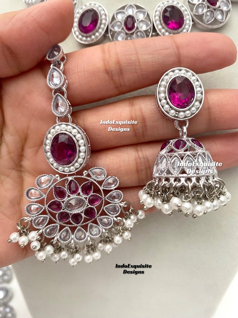 Op de afbeelding: Een zilveren ketting en oorbellen set met roze en witte stenen. De ketting heeft een hanger met een rond ontwerp en de oorbellen hebben een traanvorm.