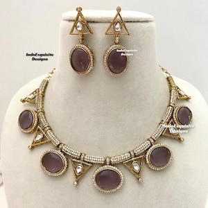 May include: A gold necklace with a geometric design and purple stones. The necklace has a row of small clear stones and a larger purple stone in the center. The necklace is paired with matching earrings with a similar design.