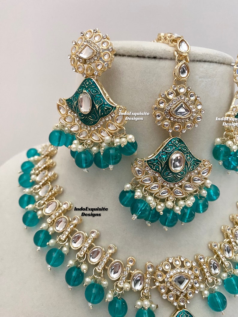 May include: A gold and green Indian bridal necklace set with teardrop-shaped pendants, clear stones, and green glass beads. The set includes a necklace, earrings, and a maang tikka.