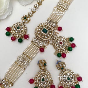 May include: An elaborate jewellery set comprising a necklace, earrings, and a headpiece. The set features faux pearls, gold-toned metal, and green and red bead accents. The design showcases intricate detailing and a traditional style, ideal for special events.