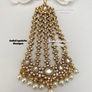 May include: Gold and white Indian bridal hair accessory with pearl drops. The accessory is made of metal and has a floral design. It is called a "maang tikka" and is worn on the forehead.