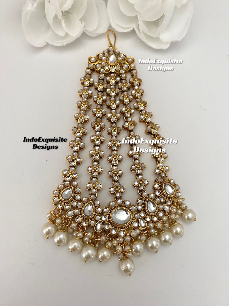 May include: Gold and white Indian bridal hair accessory with pearl drops. The accessory is made of metal and has a floral design. It is called a "maang tikka" and is worn on the forehead.