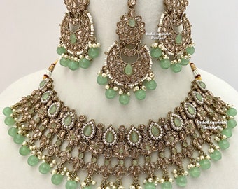 Puede incluir: Un conjunto de collar de novia indio en oro y verde que incluye un collar gargantilla, pendientes y un maang tikka. El conjunto presenta intrincados diseños florales con piedras transparentes y rosas, y cuentas verdes.