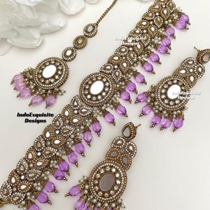 May include: A gold-tone Indian bridal jewelry set with purple glass beads and mirror accents. The set includes a necklace, a maang tikka, and earrings.