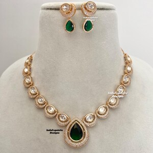 May include: A gold necklace and earrings set featuring a teardrop-shaped green stone surrounded by clear stones. The necklace has a row of clear stones around the teardrop stone. The earrings are crescent-shaped with a clear stone and a teardrop-shaped green stone hanging from the bottom. The text "IndoExquisite Designs" appears on the image.