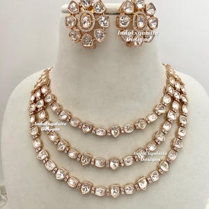 Puede incluir: Un collar de oro de tres hileras con piedras transparentes y facetadas. El collar se combina con pendientes a juego. Los pendientes también son de oro y presentan piedras transparentes y facetadas. El texto "IndoExquisite Designs" es visible en la imagen.