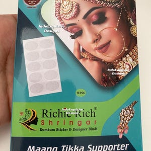 May include: A box of 'Richie Rich Shringar' Maang Tikka Supporter. The box is teal and green with a picture of a woman wearing traditional Indian jewelry. The box also contains 10 pieces of Kumkum stickers.