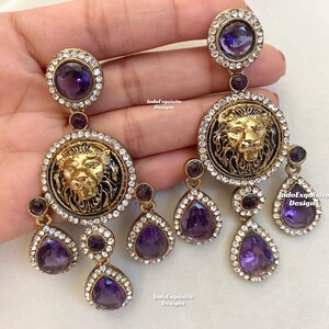 May include: A pair of gold-tone earrings with a lion head design and purple teardrop-shaped crystals. The earrings are dangling and have a total of five crystals each.