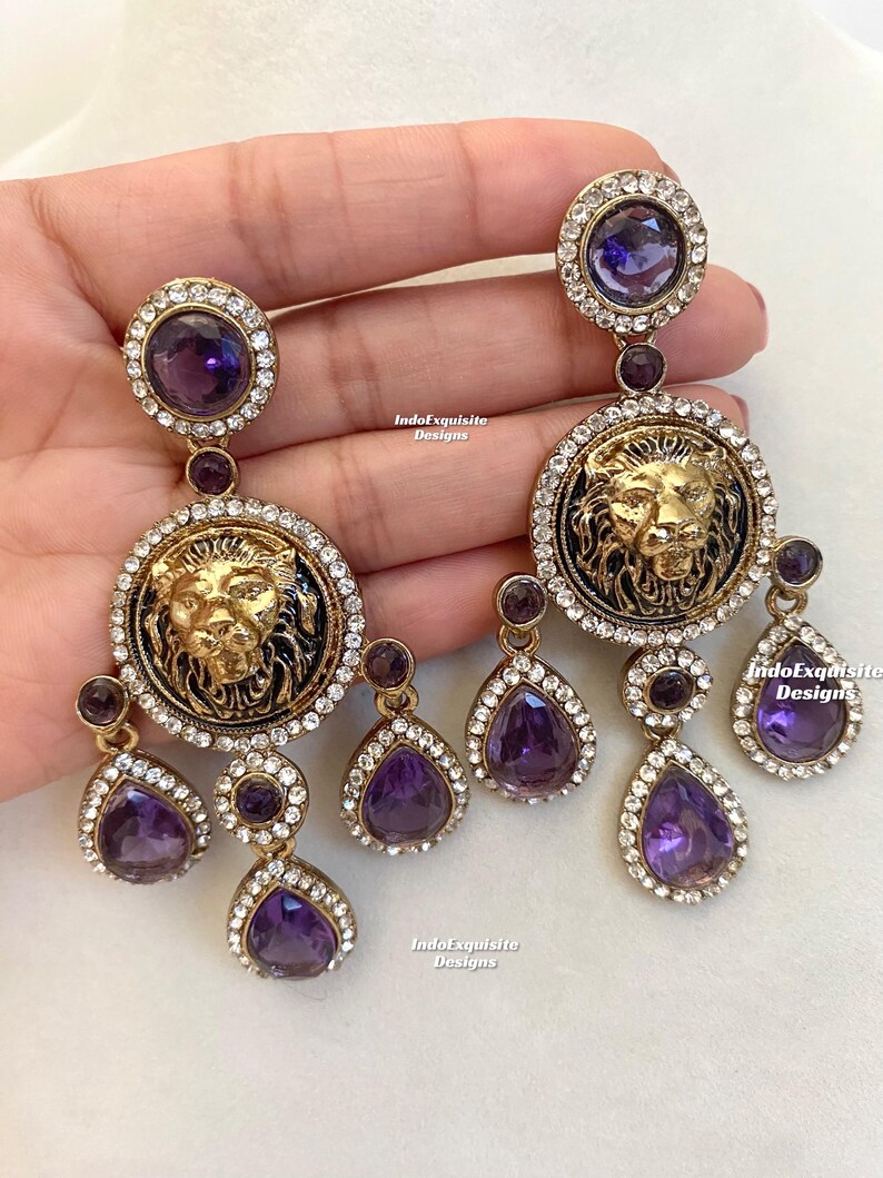 May include: A pair of gold-tone earrings with a lion head design and purple teardrop-shaped crystals. The earrings are dangling and have a total of five crystals each.
