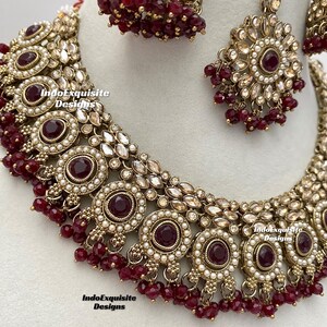 May include: A maroon and gold Indian bridal necklace set with pearl and rhinestone accents. The set includes a choker necklace, earrings, and a maang tikka.