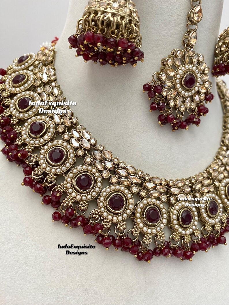 May include: A maroon and gold Indian bridal necklace set with pearl and rhinestone accents. The set includes a choker necklace, earrings, and a maang tikka.