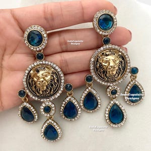 May include: A pair of gold-tone earrings with a lion head design and blue crystal accents. The earrings feature a large round pendant with a lion head in the center, surrounded by smaller blue crystals. The lion head is detailed with a mane and a crown. The earrings are suspended from a smaller blue crystal drop.